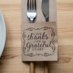 Top view of metal fork and knife in paper cover decorated with Give Thanks With A Grateful Heart inscription placed on wooden table